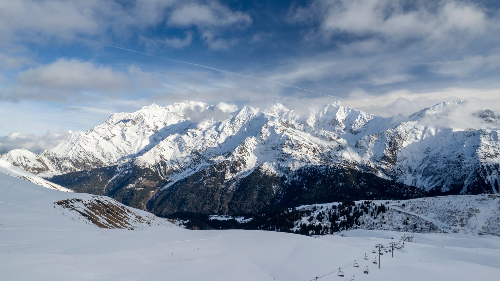 Snow-covered Alpine peaks under a blue sky, a chairlift climbing the pisted slope in the foreground